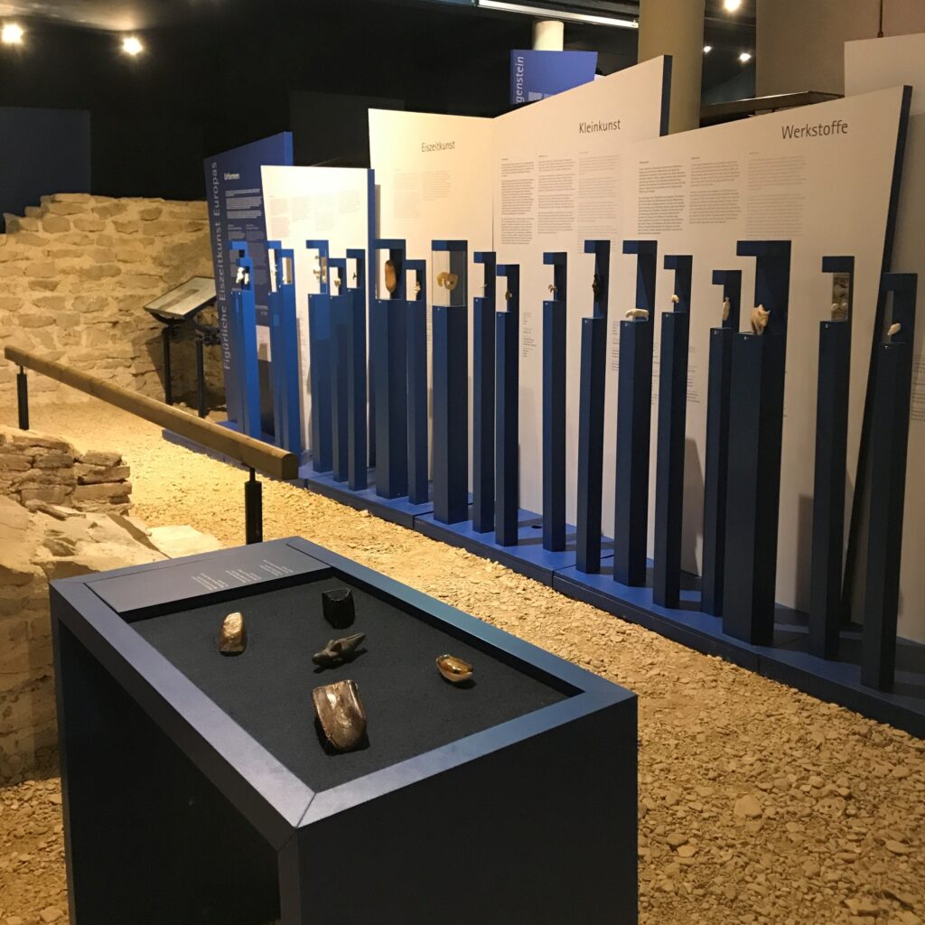 Ausstellung Urformen und Eiszeitkunst mit archäologischen Fundstücken im Museum im Römerbad: Im Vordergrund ist ein Tisch mit Materialen aus der Steinzeit zum berühren, im Hintergrund aufgereihte blaue Sockel mit Exponaten und Informationstafeln zu Eiszeitkunst, Kleinkunst und Werkstoffen. Links ein freigelegter Mauerrest des Römerbades auf Schotterboden