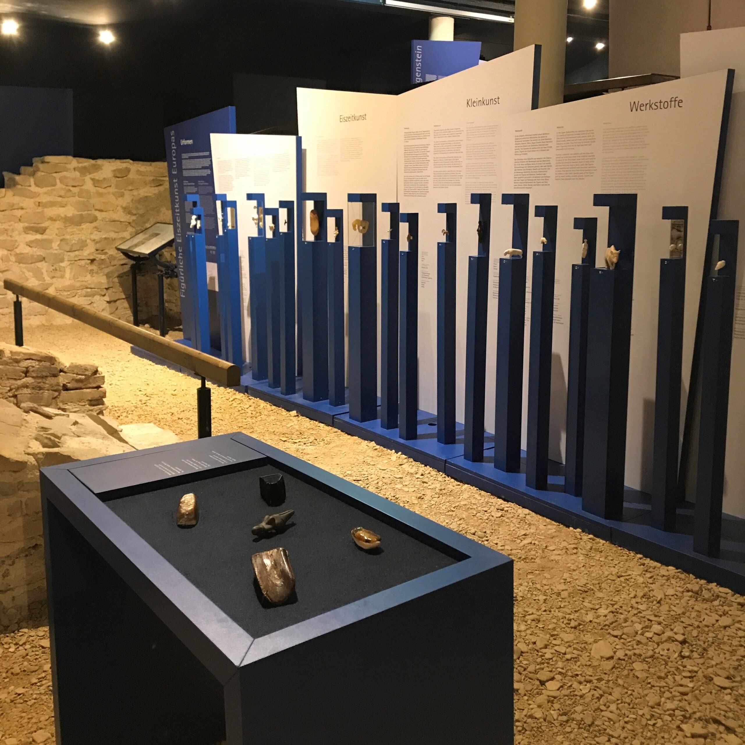 Ausstellung Urformen und Eiszeitkunst mit archäologischen Fundstücken im Museum im Römerbad: Im Vordergrund ist ein Tisch mit Materialen aus der Steinzeit zum berühren, im Hintergrund aufgereihte blaue Sockel mit Exponaten und Informationstafeln zu Eiszeitkunst, Kleinkunst und Werkstoffen. Links ein freigelegter Mauerrest des Römerbades auf Schotterboden