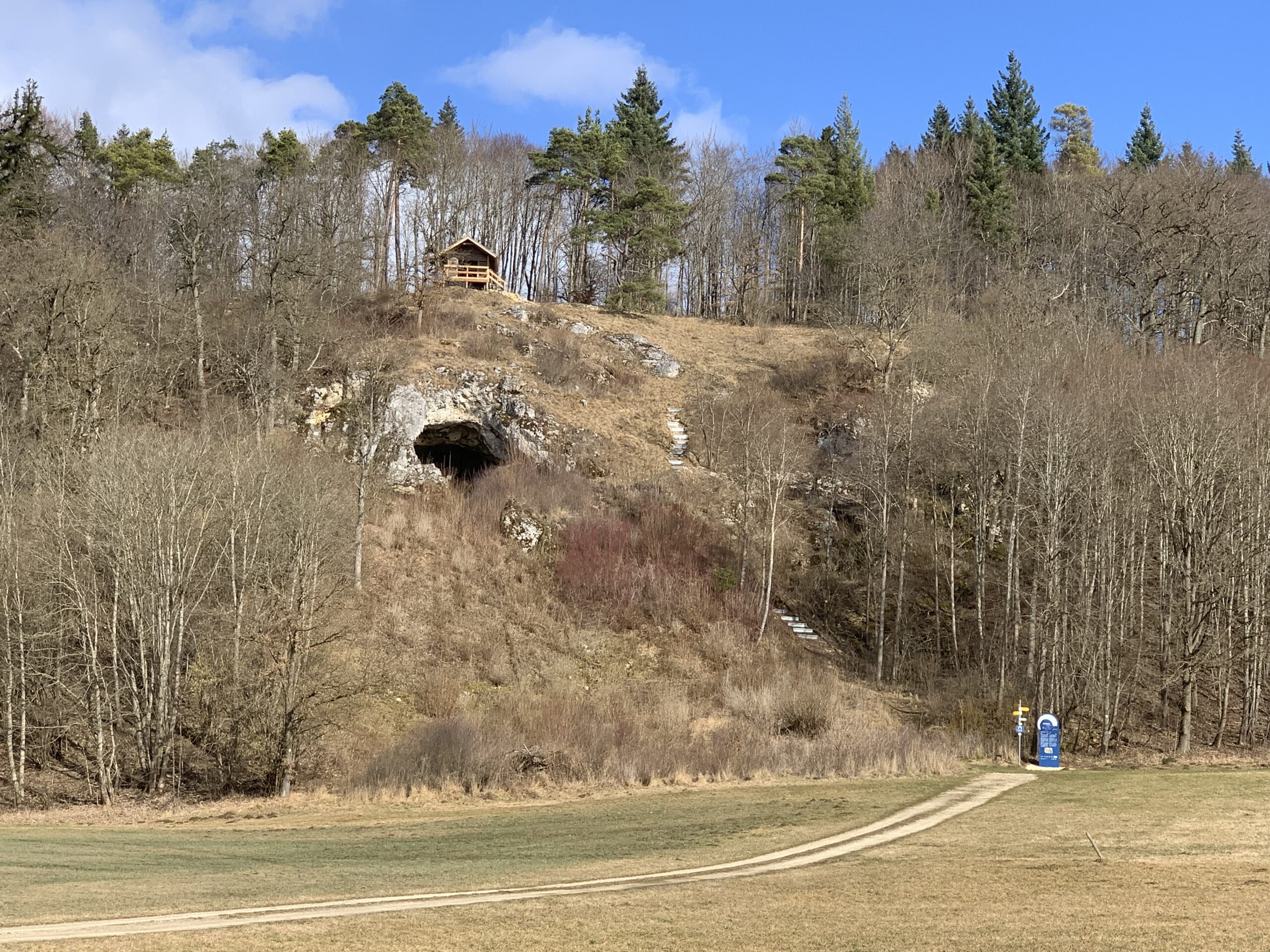 Blick auf Bocksteinhöhle in einem bewaldeten Hügel. Am Fuße des Hügels verläuft ein schmaler Weg, auf dem ein blaues Informationsschild steht. Eine Treppe führt von unten zur Höhle, die in einen Felsen hineinführt. Oberhalb der Höhle befindet sich ein Schutzhäuschen, das auf den Hügel gebaut wurde. Der Himmel ist klar, und die Bäume sind noch winterlich kahl.