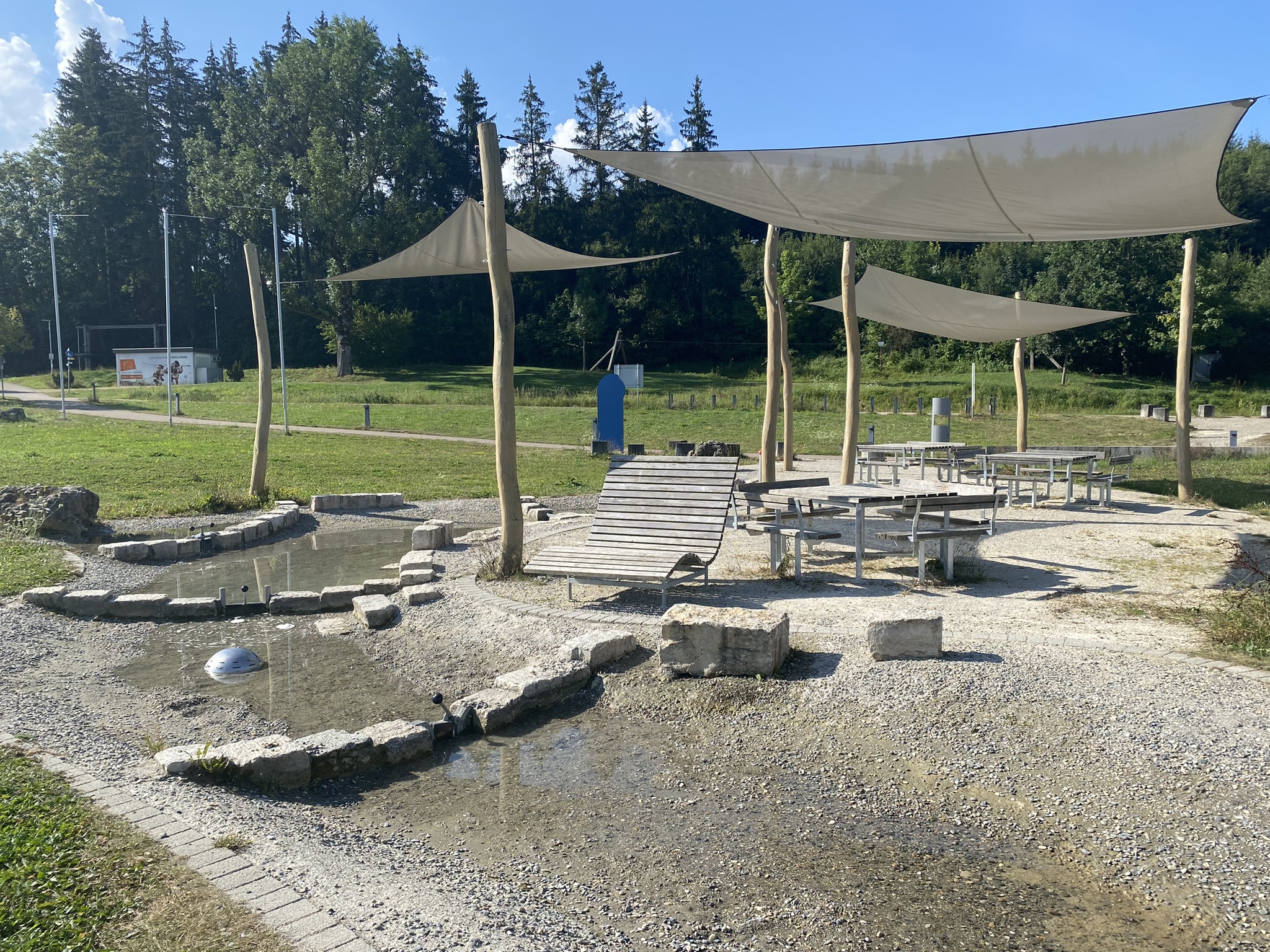 Wasserspielplatz mit Schotter- und Sandflächen, Holzliegen, Tischen und großen Sonnensegeln in einer grünen Parklandschaft