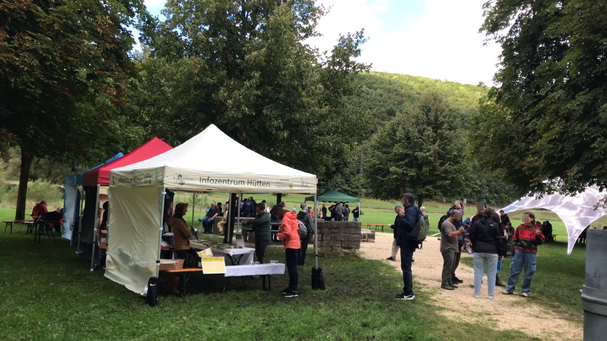 Mehrere Menschen stehen und sitzen bei mehreren Informationsständen mit bunten Zelten und Tischen an einer Freifläche mit Bäumen und Hügeln im Hintergrund