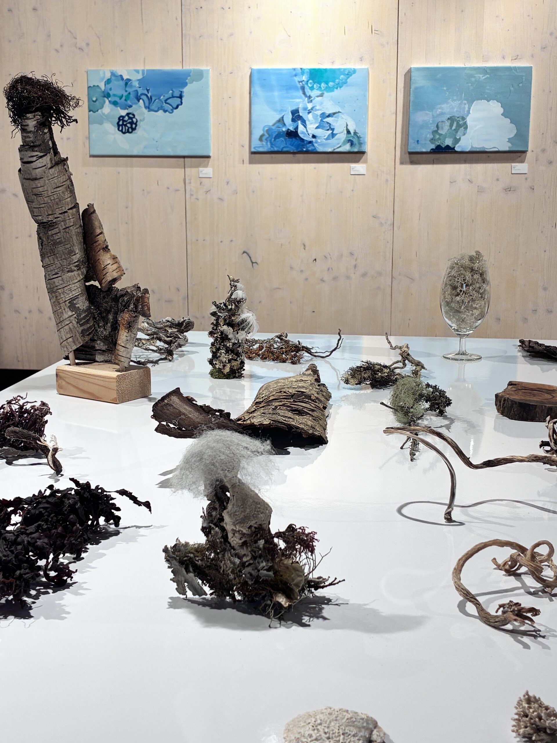Kunstinstallation mit verschiedenen organischen und natürlichen Materialien auf einem weißen Tisch vor einer Holzwand mit drei abstrakten blauen Gemälden